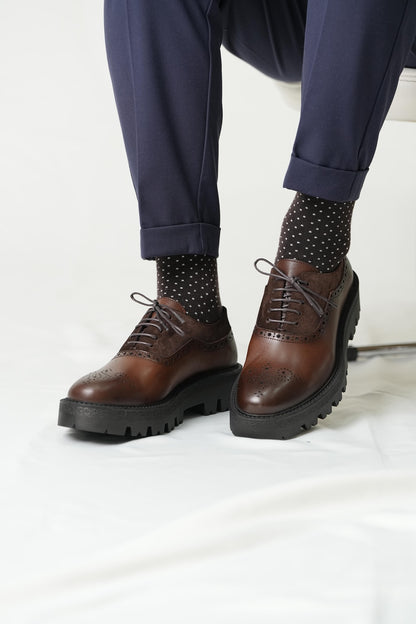Brown leather shoes with thick soles worn with navy pants and polka dot socks on a white background