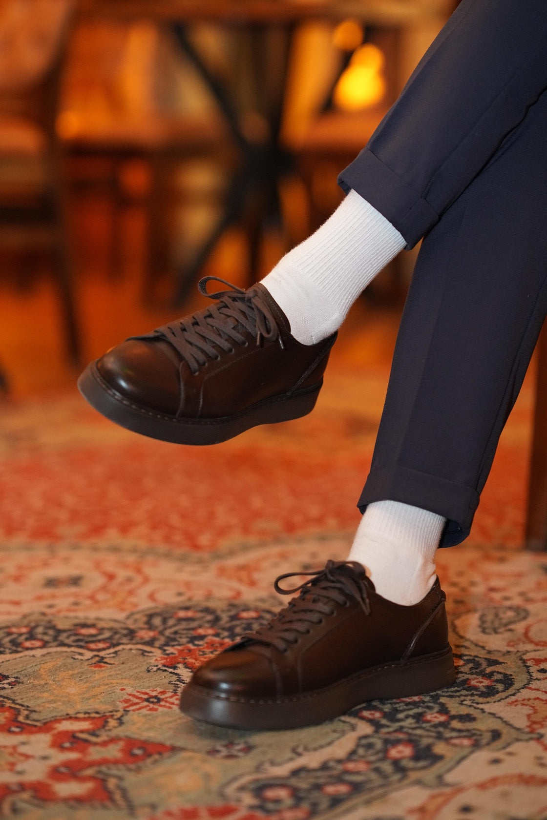 Brown leather shoes sneakers worn with dark pants and white socks on a patterned rug.